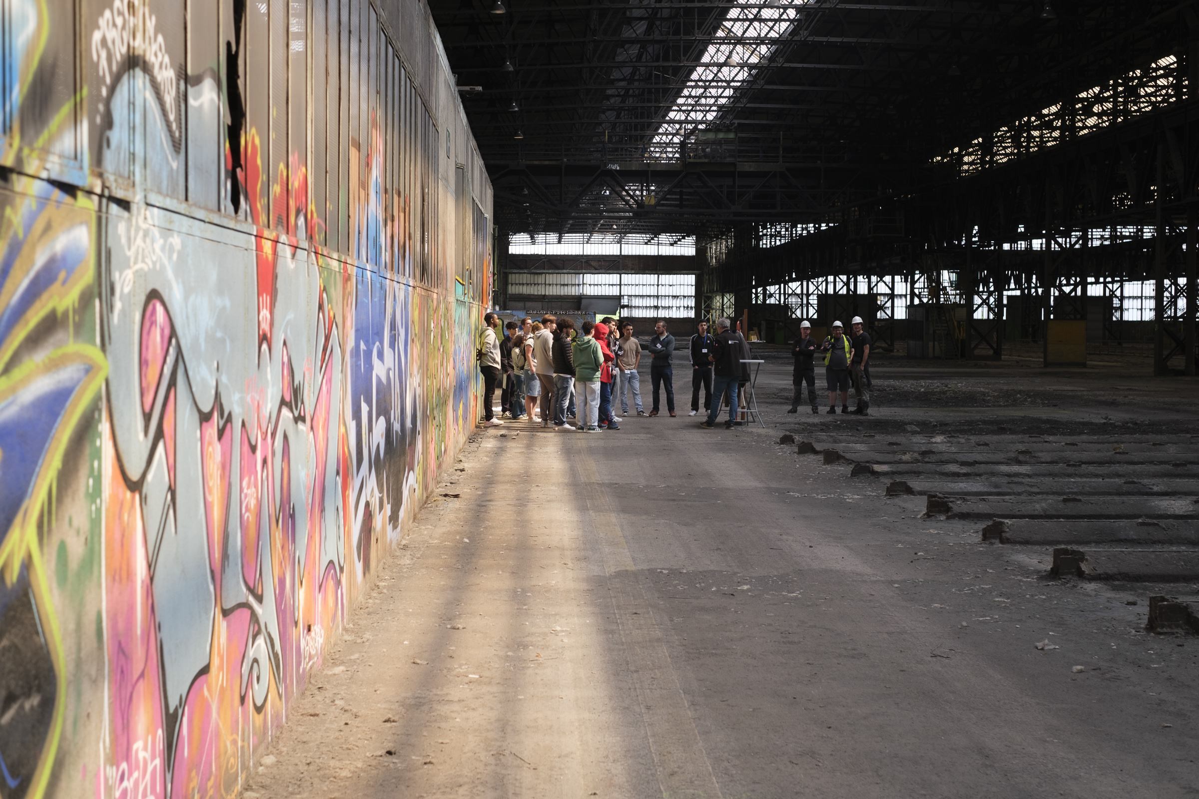 Eine Gruppe aus Jugendlichen und Arbeitern vor bunter Wand in Industriehalle.